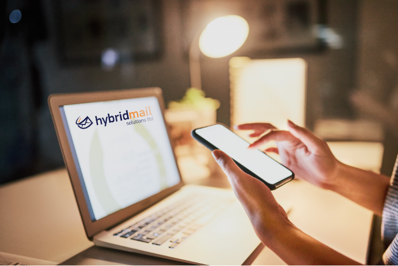 Person using a smartphone while working on a laptop displaying the Hybrid Mail Solutions logo, representing digital communication and social media strategy.